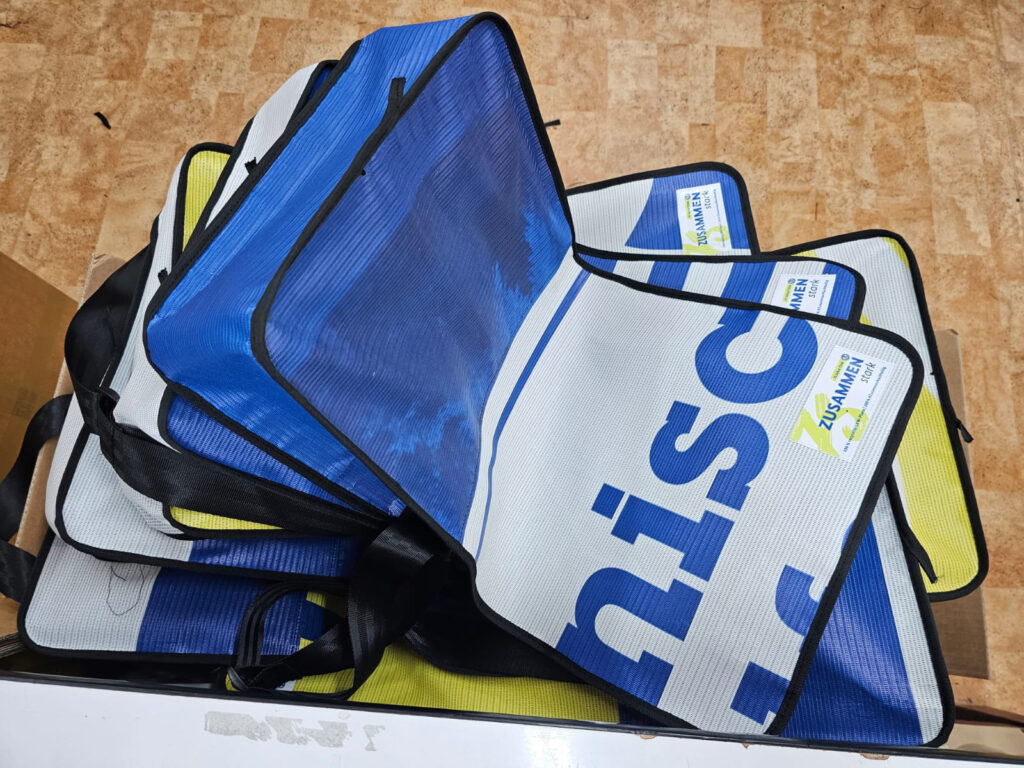 Fertige Messenger-Taschen in Blau, Weiss und Gelb gestapelt, jede mit anderem Postermotiv