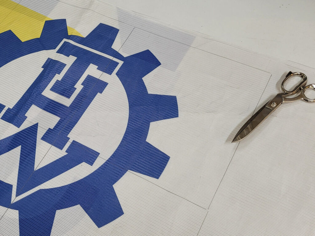 Riesenposter mit dem blauen THW-Zahnrad-Logo auf dem Zuschnitttisch, daneben die Schneiderschere